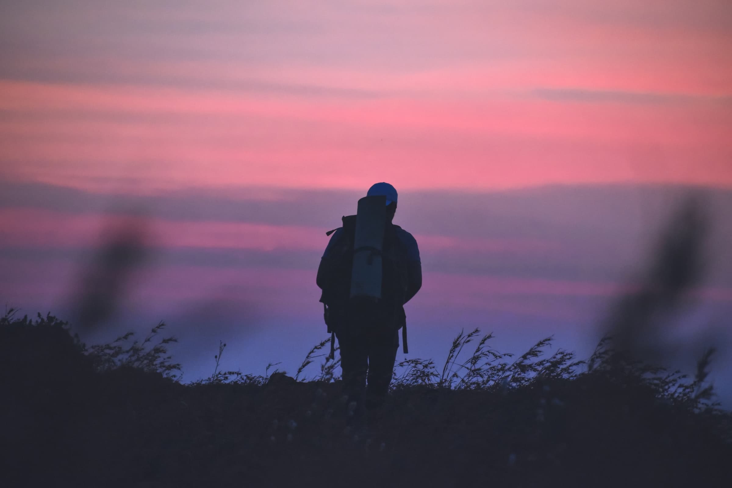 A man wearing a black jacket and backpack standing on a grass field during sunset